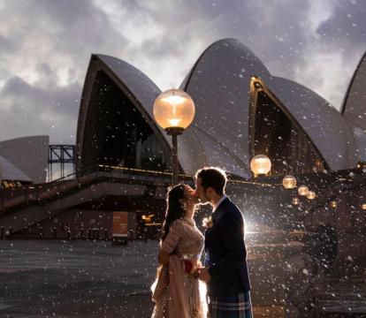 A woman in a wedding dress and a man in a suit kissing outside of the Sydney Opera House.