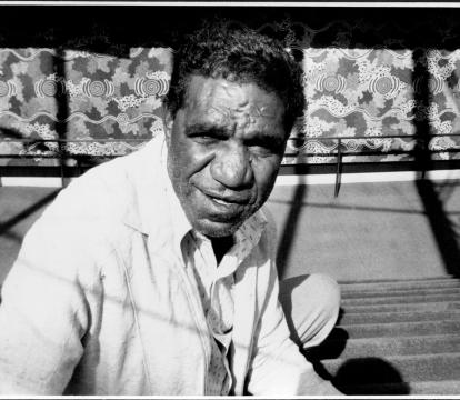 Jagamara photographed in black and white sitting on carpeted steps in front of the mural 'Possum Dreaming'.