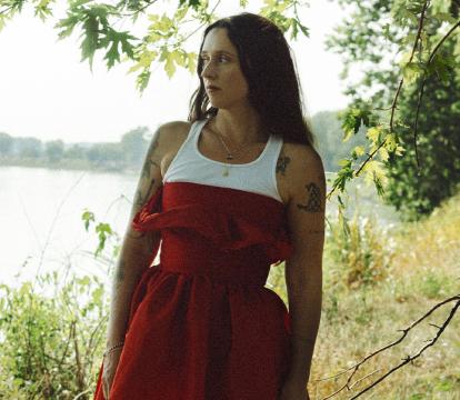 Woman in red dress standing by river, gazing into distance.