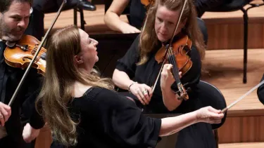 A conductor leads an orchestra, gesturing with a baton, while musicians around her play violins.