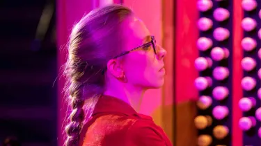 A woman with glasses and a long braid, wearing a red shirt, is illuminated by pink and purple light while playing a pipe organ.