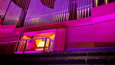 A large pipe organ illuminated by purple and pink lights, with a person seated at the organ console.