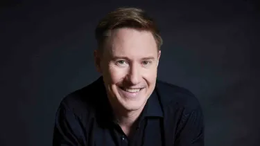 A smiling man with light brown hair wearing a dark shirt sits against a dark, softly lit background.