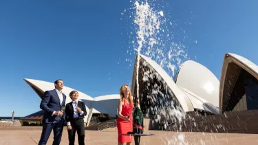 A woman in a red dress and two men in suit standing outside the Sydney opera House, watching a bottle of Champagne spray into the air.