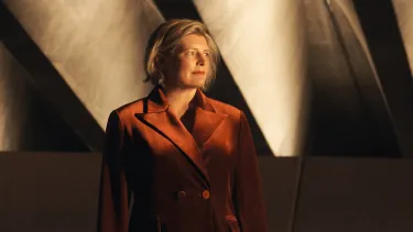 A woman wearing a dark orange jacket looking off to the side, with concrete beams in the background.