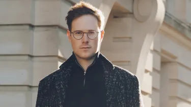 A portrait of a man with glasses in a dark shirt and coat standing in front of a stone building on a sunny day.