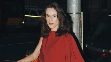 A woman wearing a red asymmetrical dress outside at night with the back of a dark grey car in the background.