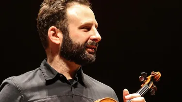 A cropped portrait of a man holding a violin against a dark backdrop.