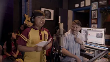 A man smiling and a child in red school uniform singing in a recording studio.