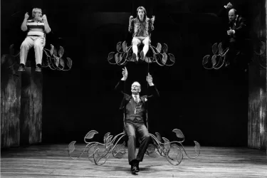 Actors on stage, three of them seated above a man.