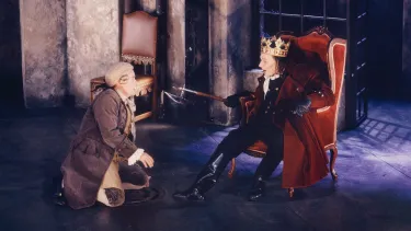 An actor in a blonde wig and brown coat kneeling in front of a king.