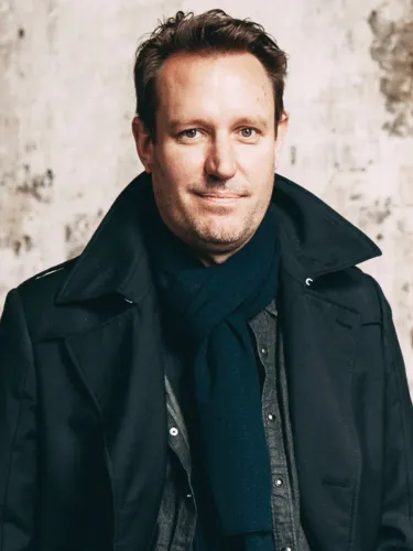 A middle aged man wearing a long black coat. and a scarf.
