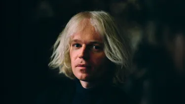 A white man in early 40s with jaw-length white hair looks at the camera.