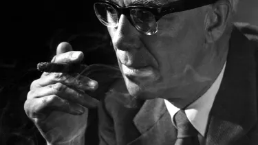 A person wearing glasses, in a suit and tie smoking a cigar.