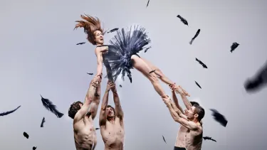 A performer in a tutu is held in the air by 4 other performers against a light grey background with feathers floating in the foreground and background.
