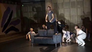 The cast gather around a performer standing on the couch.