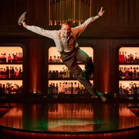 A lively performer in vintage attire jumps energetically on a stage, with shelves of colorful bottles in the background. The scene is vibrant and dynamic.