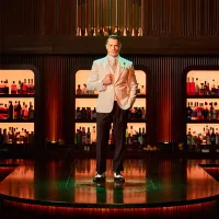 A man in a white suit stands confidently in front of a well-lit bar, filled with assorted bottles and warm wood tones, exuding an elegant, upscale ambiance.