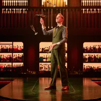 A man in a green suit and bow tie balances a tray while juggling champagne flutes in a sophisticated, warmly-lit bar stocked with various bottles.