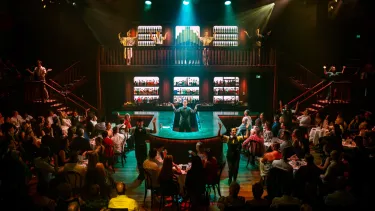 A full theatre with a green light cast on the stage. Dancers are mid routine with their arms in the air.