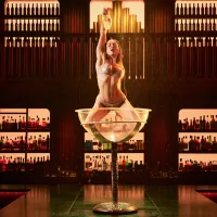 A performer in a silver bikini poses confidently in a giant champagne glass, holding a bottle aloft. The background features shelves of backlit bottles.