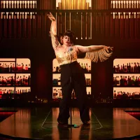 Performer in a brimmed hat and fringed gold top poses energetically on a stage.