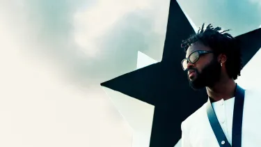 A young black man wears sunglasses and poses in front of a large black star.