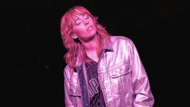 A white woman in her 30s wears a pink reflective jacket and meditatively closes her eyes. She's bathed in pink light.