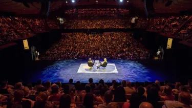 Two women sit on the Concert Hall stage in conversation to a full audience with a timer in front of them.