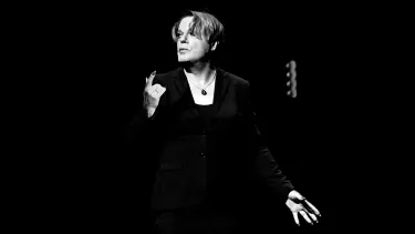 A person in a dark suit gestures passionately onstage in a spotlight against a black background, conveying intensity and focus.