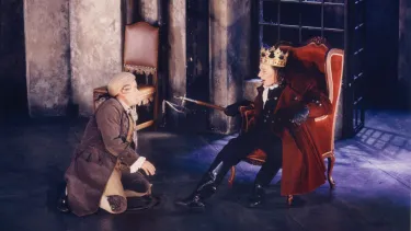 An actor in a blonde wig and brown coat kneeling in front of a king.