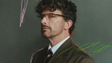 A man wearing glasses and a dark green suit looking sidelong against a grey-green background with subtle scribbled marks in pink, red and green.