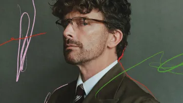 A man wearing glasses and a dark green suit looking sidelong against a grey-green background with subtle scribbled marks in pink, red and green.