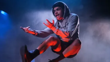 A boy in a hoodie and shorts performs a high jump on stage, arms expressive. Blue and red lights create a dynamic and dramatic atmosphere.