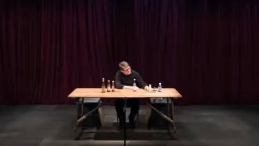 A person sits alone at a wooden table in a dimly lit room with deep red curtains. Bottles are organized on shelves against the walls, creating an intimate, contemplative mood.