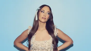 A white woman wears a blue bow in her long brown hair posing with her hands on her hips.