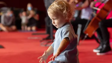 A toddler in a blue sweater and floral skirt dances on a red carpeted floor with people and musicians playing string instruments in the background.