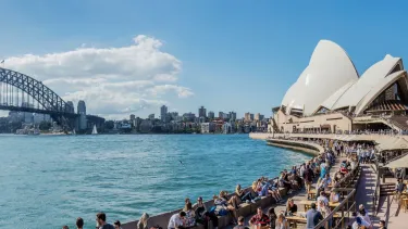 Panoramic view of Sydney Harbour with the Sydney Opera House on the right and Sydney Harbour Bridge on the left, with numerous people and waterfront restaurants in the foreground.