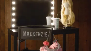 A long blonde wig and cup with a straw sit on a black dressing table with a mirror edged with lights and a folding chair with Mackenzie on the back and a pink handbag hanging off one side.