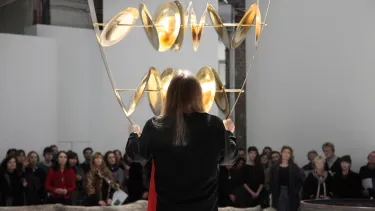 A perfomer stands in front of a large sculpture of suspended gold plates in an art gallery.