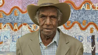 Michael Nelson Jagamara wearing a beige suit jacket over a blue and white checked shirt and wide brim hat, standing in front of an artwork in pastel colours.