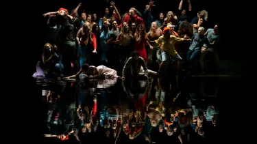 A large group of people in colourful clothing pose infront of a body of water with a black backdrop.