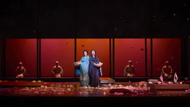 Two women sing with bowls in their hands in a red backdrop.