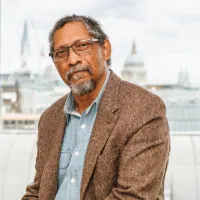 A black man stares at the camera, he wears black glasses and has a beard and grey hair.