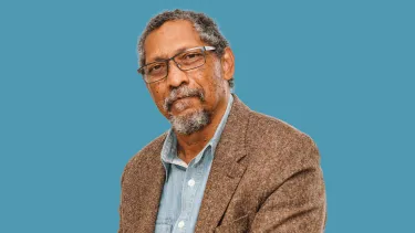 A black man stares at the camera, he wears black glasses and has a beard and grey hair.