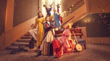 Five women stand staggered on a staircase in regency era gowns, with musical instruments for Pride and Prejudice (sort of) as confetti falls around them.