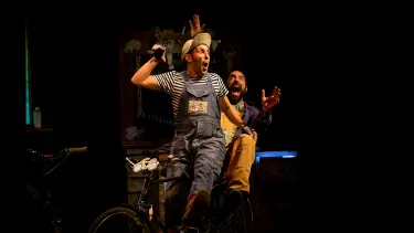 A man wearing overalls sits on a bike with a surprised, but joyful look on his face. A man appears behind him cheering with his hands out.