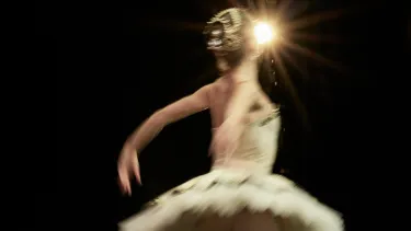 A ballerina wears a gold tutu and hairpiece. Her back is to the camera and her arms are stretched behind her, mid-dance move. A spotlight is seen behind her.