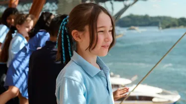 A young girl wearing a school uniform stares out a window in a Sydney Opera House foyer. The Sydney Harbour Bridge is seen in the background.