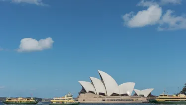 Image of the Sydney Opera House against a blue cloudy sky. The Opera House sits on the bottom of the image, with two ferries to its left, and one to its right.